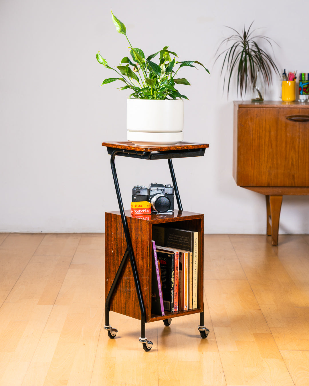 Vintage 1950s mid-century modern rolling side table, wood veneer, tubular black metal frame, chrome wheels, built-in bookcase. Vecino Shop